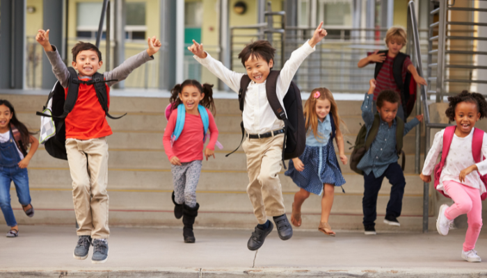 Children running and jumping outside school building with backpacks on