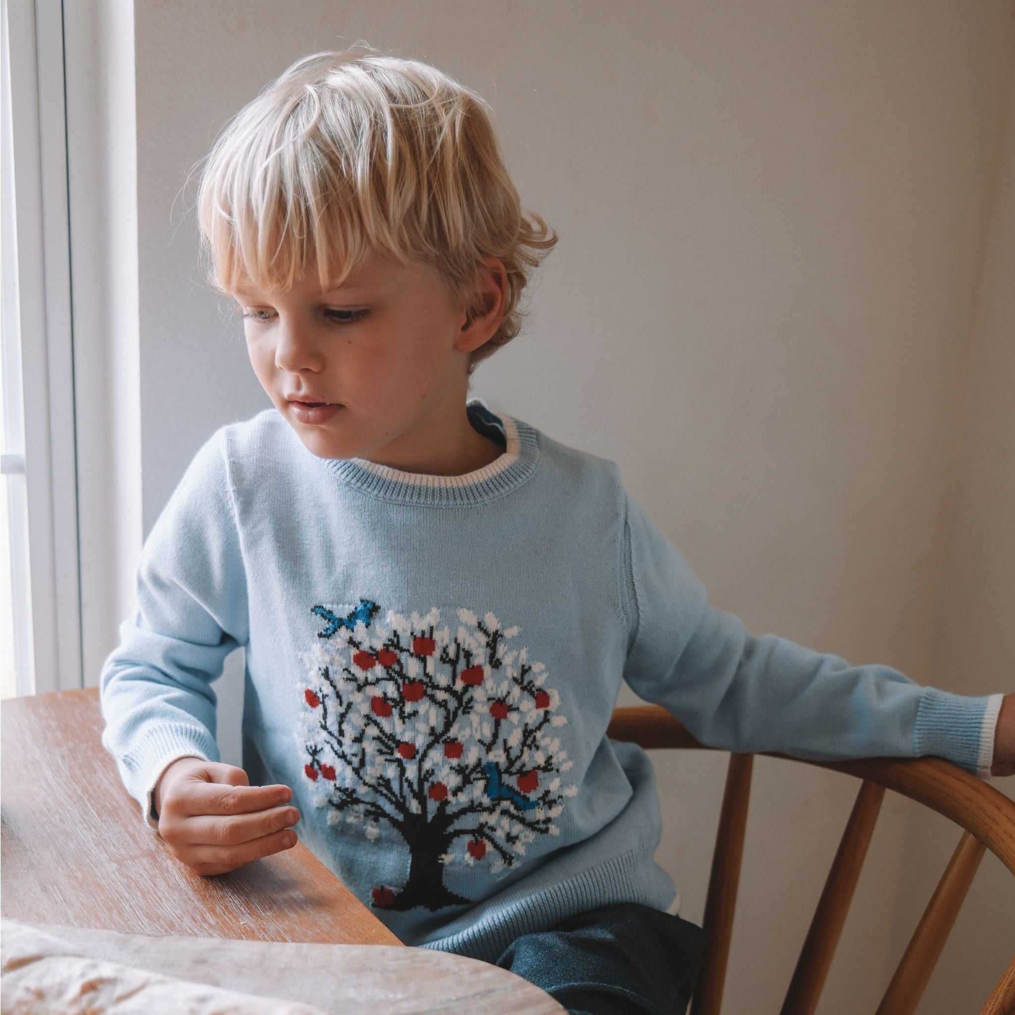 A child sitting at a table wearing a long-sleeved blue sweater with a tree intarsia design in red, white, and blue.