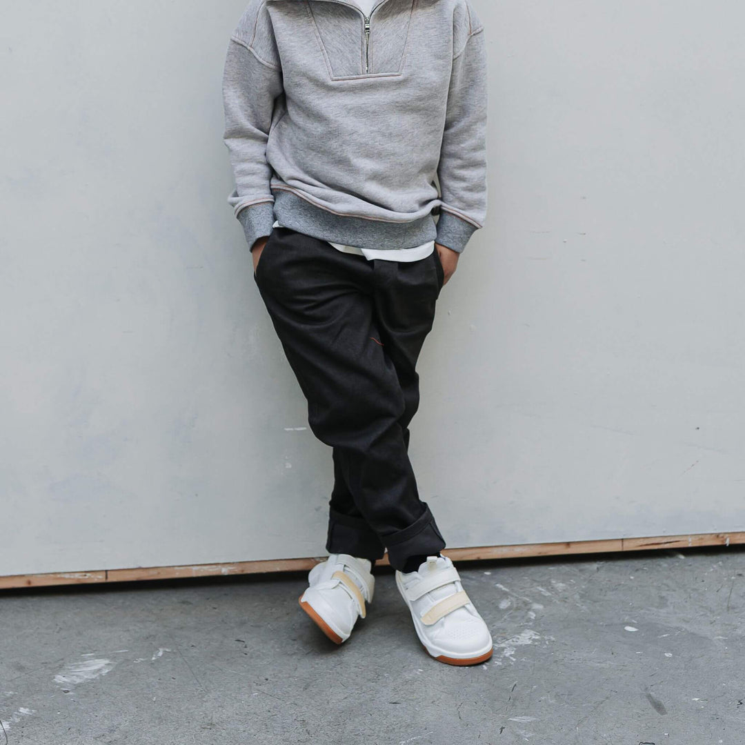 Boy in a gray half-zip sweatshirt and black Vintage Drill Pants leaning against a studio wall