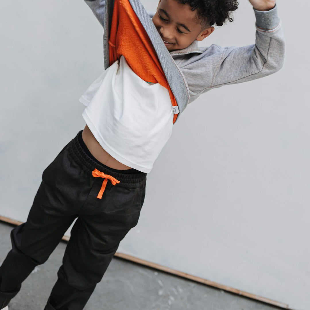 Boy pulling a sweatshirt over his head, wearing black drill pants with an orange drawstring