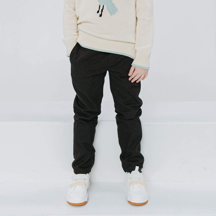 Straight-leg black drill pants on a child in a cream sweater and white sneakers, front view