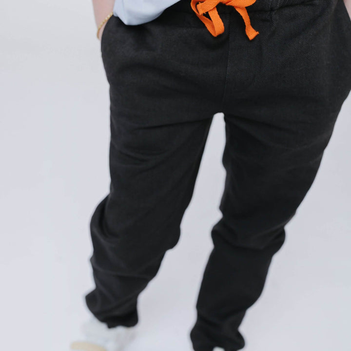 Close-up of the elastic waistband and orange drawstring on Firebird Kids' black Vintage Drill Pant, worn by a kid