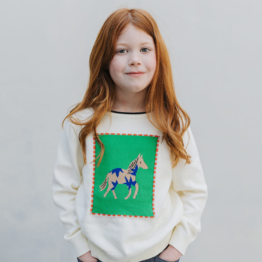Girl, waist up, in the ivory horse intarsia crewneck with green panel