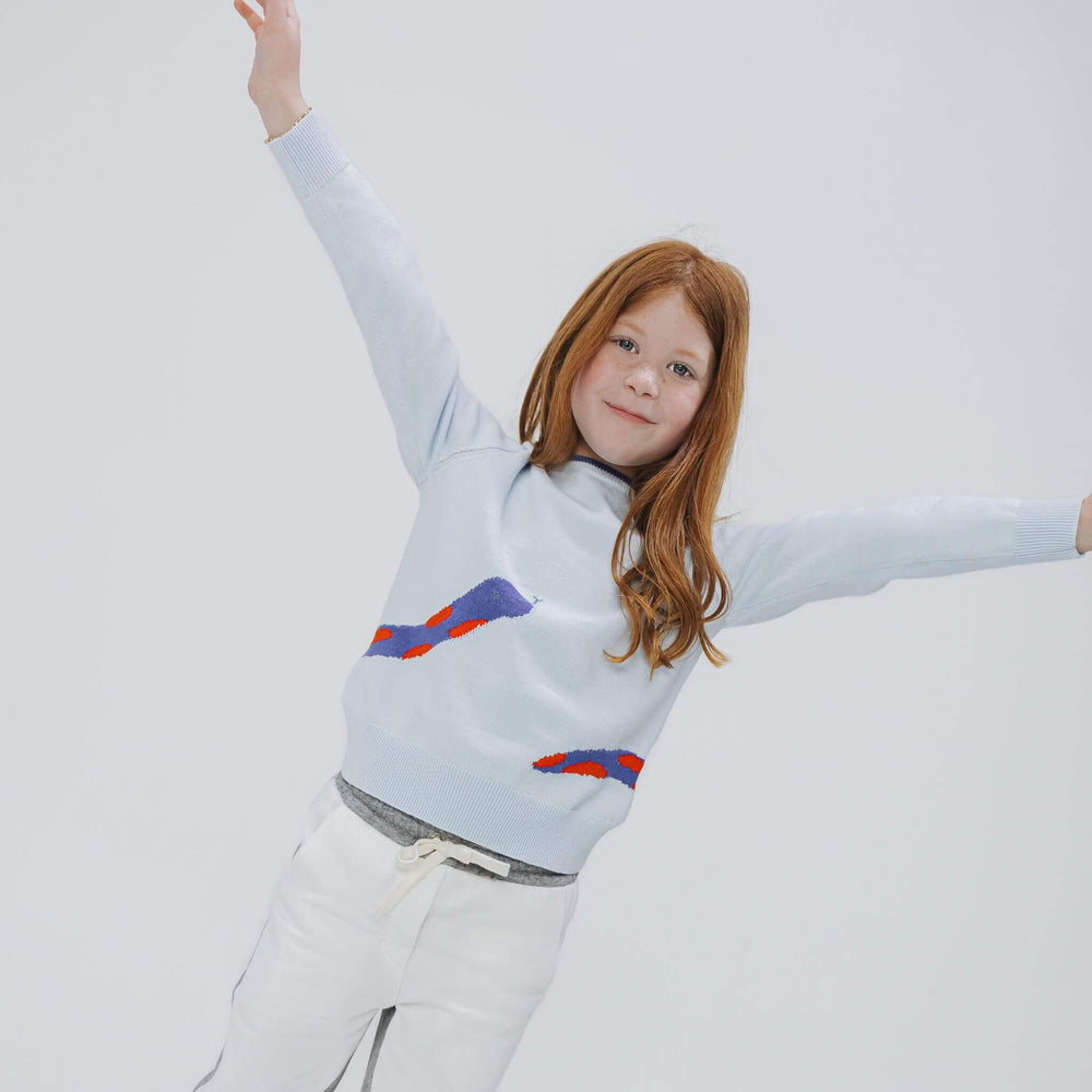 Girl jumping with arms raised in the powder blue organic cotton graphic sweater, snake wrapping toward the hem