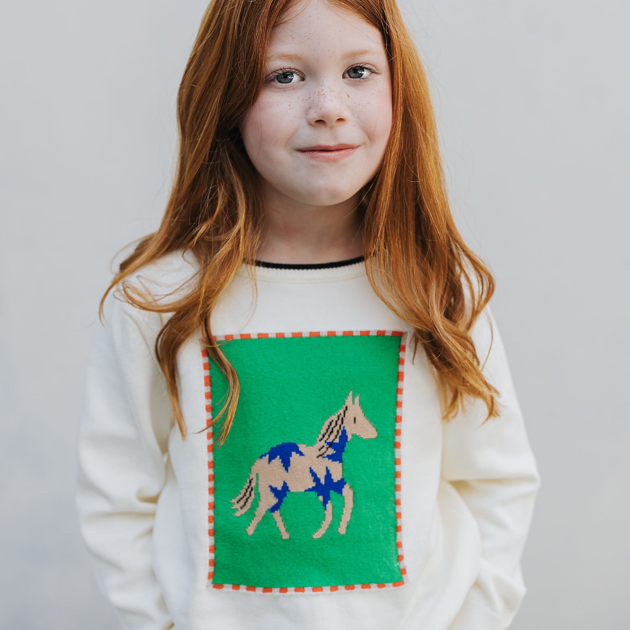 Girl wearing the ivory kids horse sweater, front view, tan and cobalt horse knitted into a kelly green panel