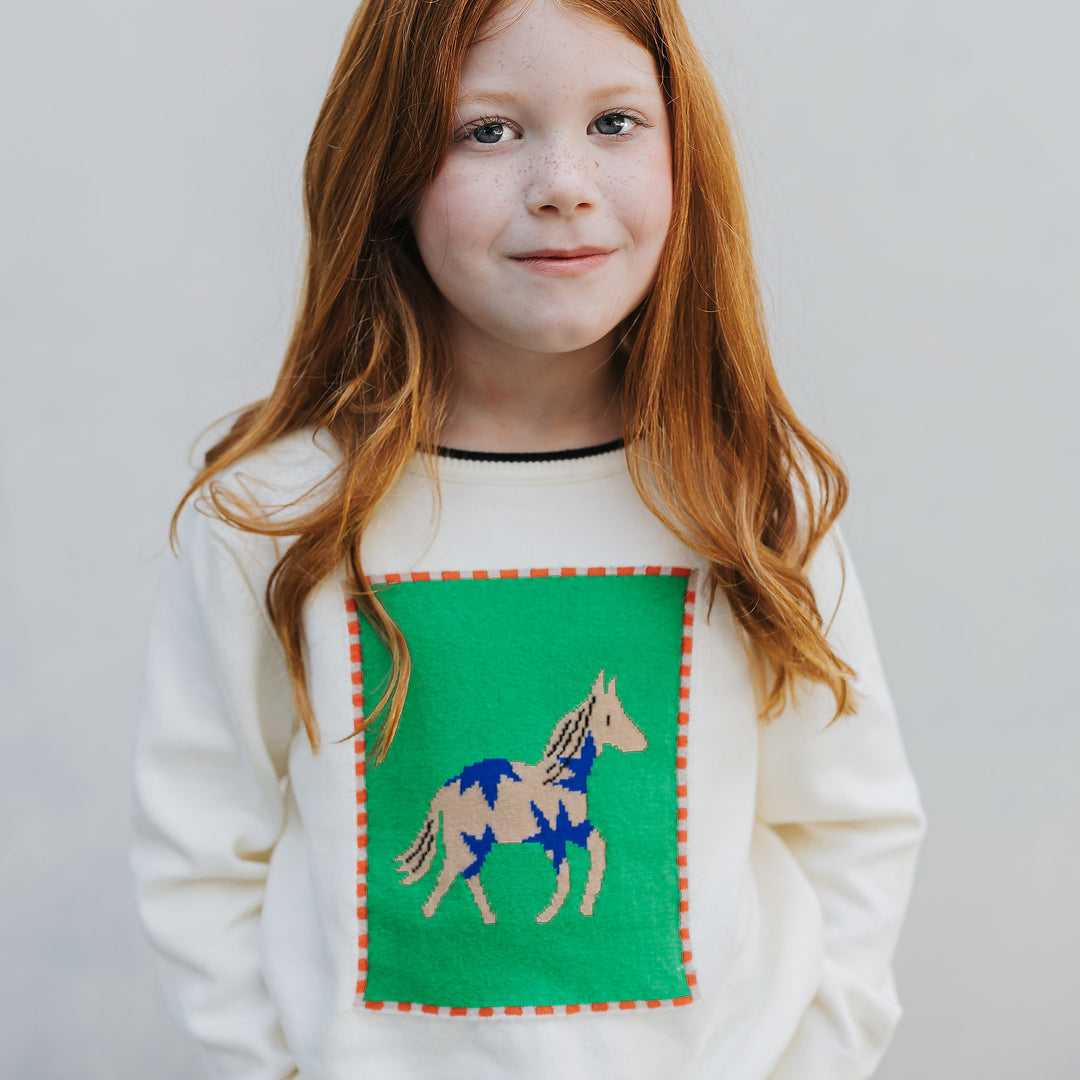 Girl wearing the ivory kids horse sweater, front view, tan and cobalt horse knitted into a kelly green panel