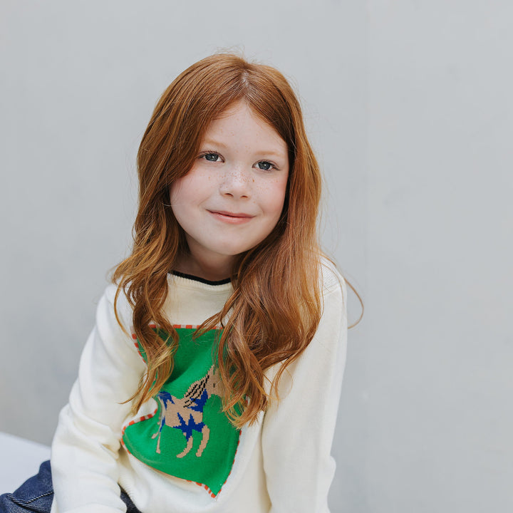 Girl seated in the ivory organic cotton horse graphic sweater